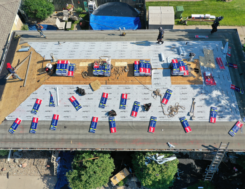 Aerial view of a flat rooftop under renovation, with workers, stacked roofing materials, and equipment scattered. Vibrant colors, sunny day.
