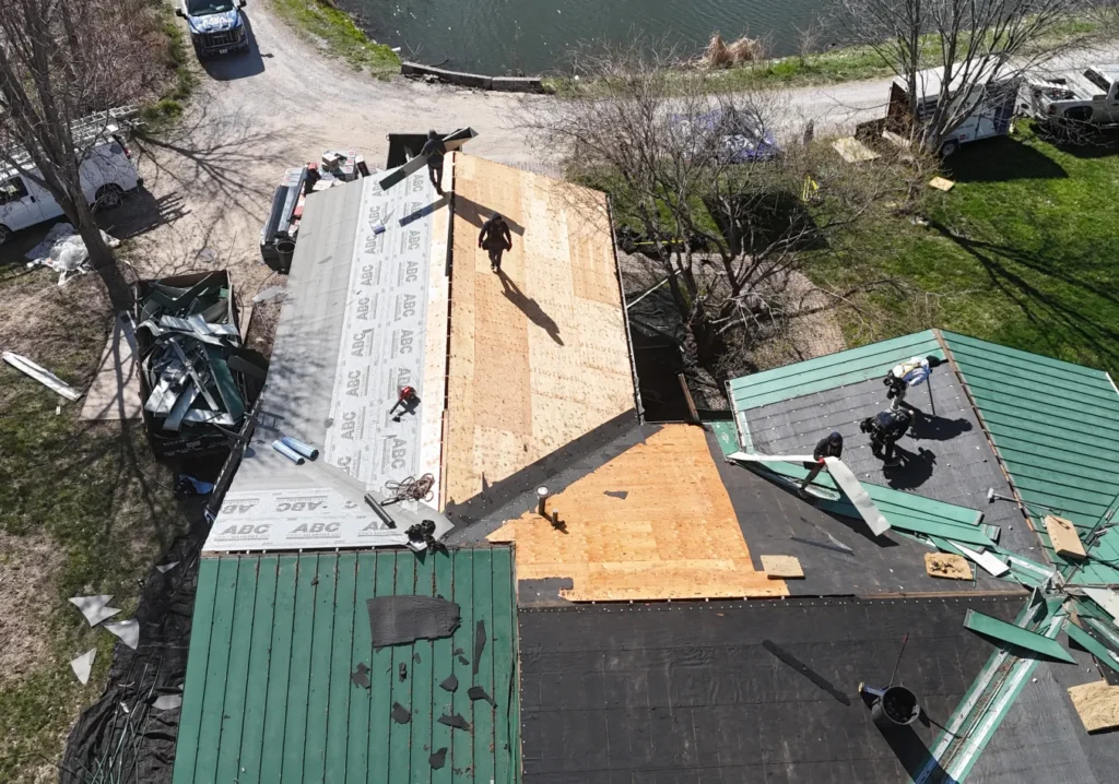Aerial view of a roof being repaired. Workers are installing new wooden panels and protective underlayment. Tools and materials are scattered around.