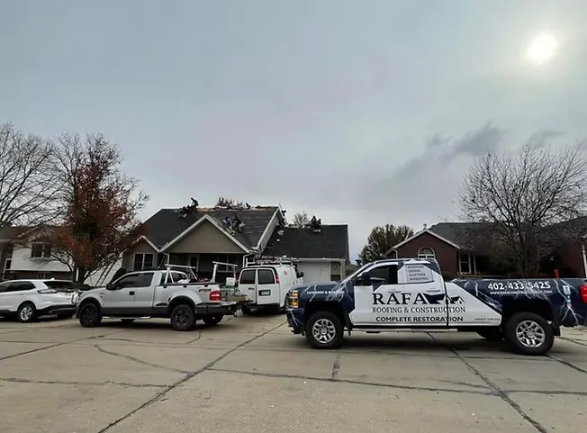 Several trucks are parked in a suburban neighborhood, with contractors working on a house roof. A truck with "Rafa Roofing & Construction" is prominently visible.