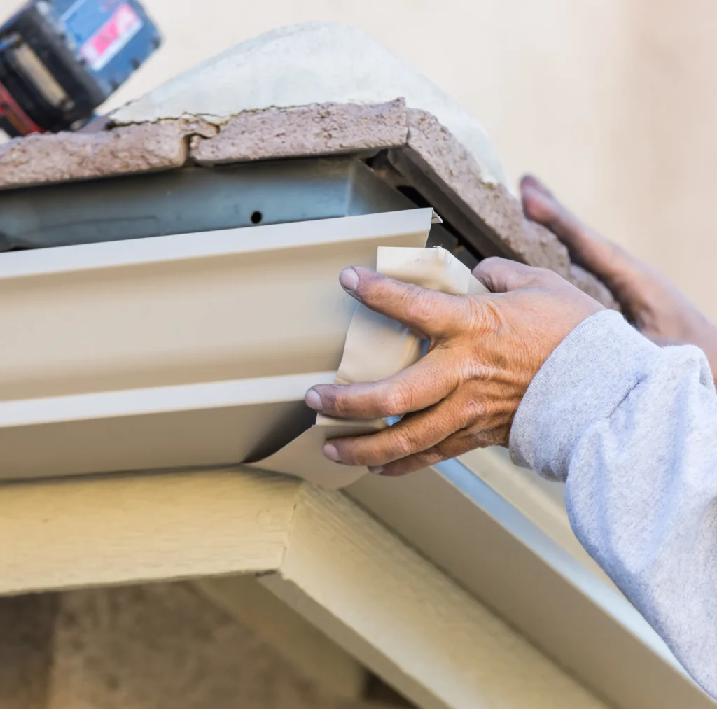 Worker in a gray shirt installs a rain gutter under a roof edge, holding it with both hands. A drill rests nearby, showing hands-on construction work.