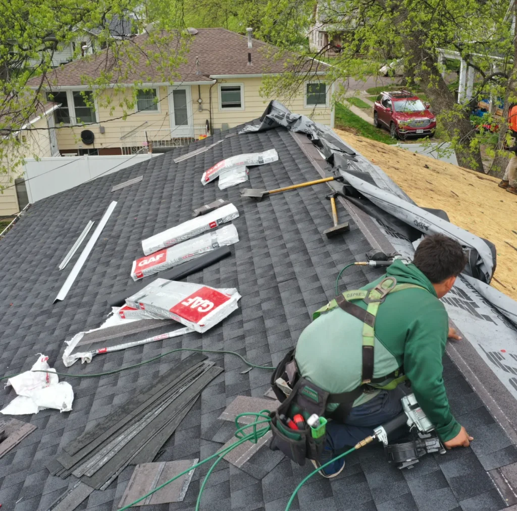 Roofer in a green hoodie kneels on a partially shingled roof, surrounded by tools and shingles. Nearby houses and trees create a suburban backdrop.