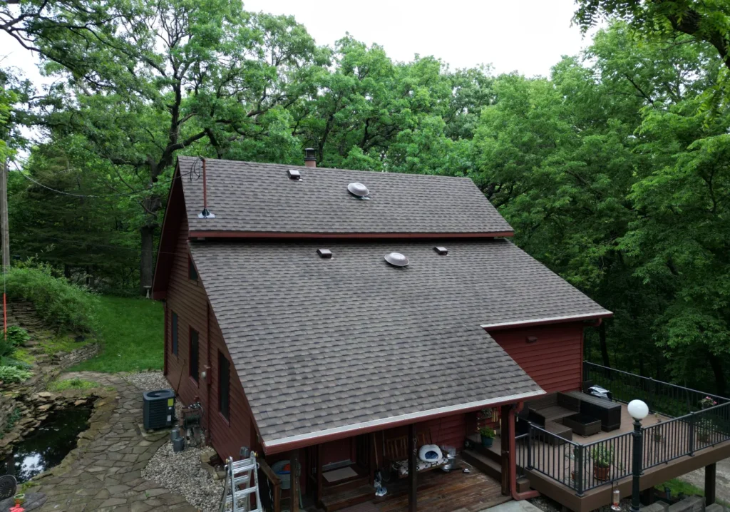A rustic red wooden house with a steep, shingled roof, nestled in lush green trees. A stone path leads to a cozy deck with outdoor furniture.