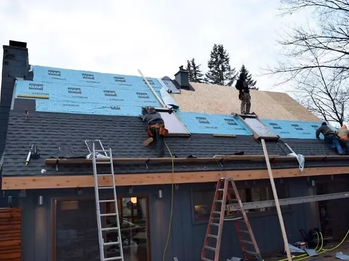 Several workers are repairing a sloped roof, with ladders and tools visible. The roof is partially covered with blue waterproof underlayment. It's a clear day.