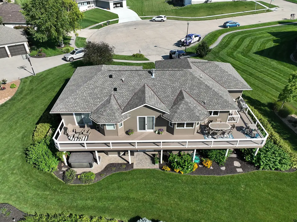 Aerial view of a suburban house with a gray roof and wooden deck, set amidst well-maintained lawns and garden beds. The scene is calm and inviting.