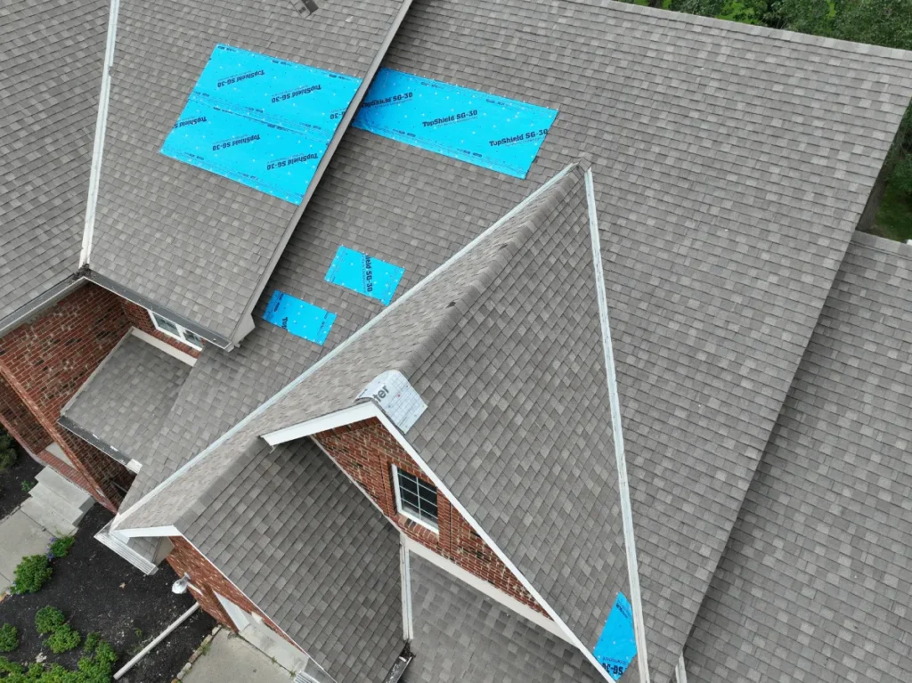 Aerial view of a brick house roof with gray shingles, showing blue tarp patches indicating repair work. The scene suggests maintenance in progress.