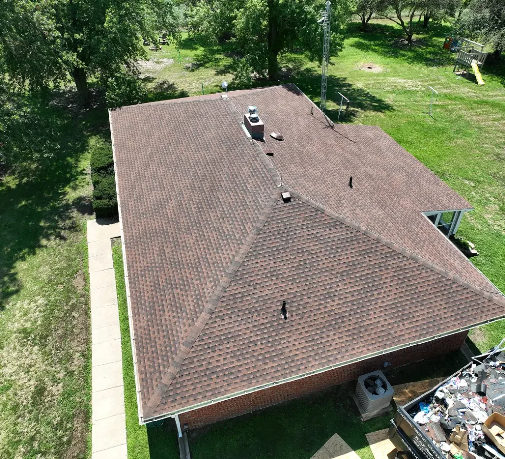 Aerial view of a house with a brown shingled roof. Surrounding greenery includes trees and grass. There's a small playground in the background.