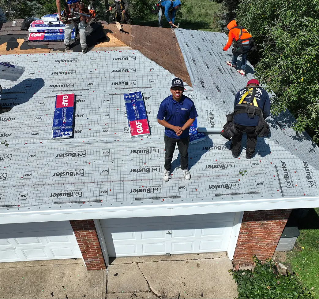Workers in safety gear install roofing shingles on a residential roof. One worker in the foreground smiles, conveying teamwork and progress.