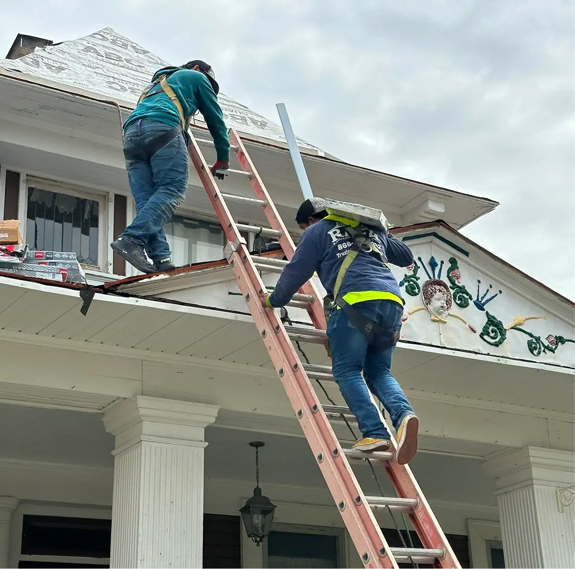Two construction workers in safety gear climb ladders to a house roof, surrounded by tools and materials, under a cloudy sky.