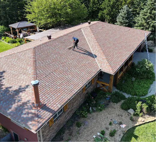 A person stands on a large, brown-shingled roof of a house surrounded by lush greenery and trees. A garden with flowerbeds borders the house.