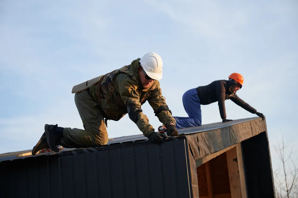 Two workers, wearing helmets and safety gear, kneel on a rooftop under clear skies, focused and intent on construction or repair work.