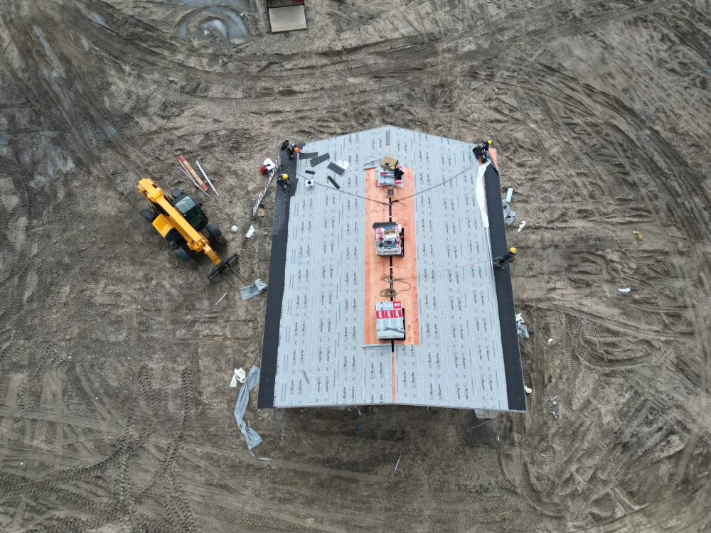 Aerial view of a construction site featuring workers installing roofing material on a large building. A yellow crane is parked on the muddy ground.