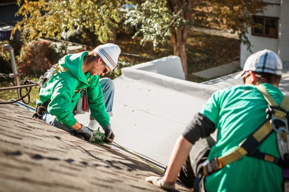 Two workers in green shirts and safety harnesses install shingles on a roof under bright sunlight, surrounded by autumn trees.