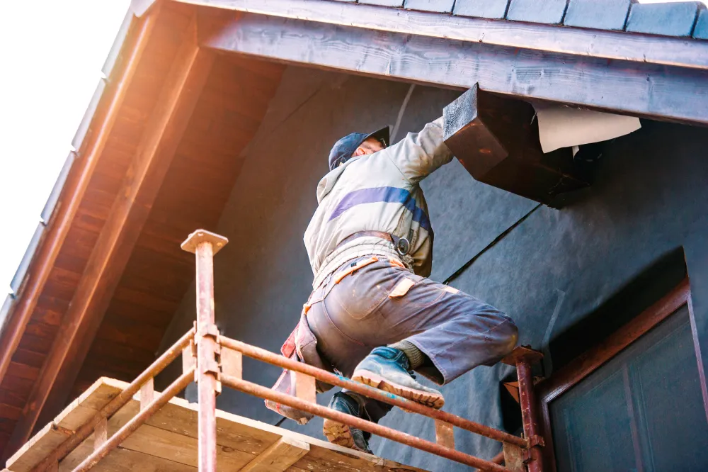 Worker on scaffolding repairs a house roof, wearing a cap and safety harness. The scene conveys diligence and focus in a construction setting.