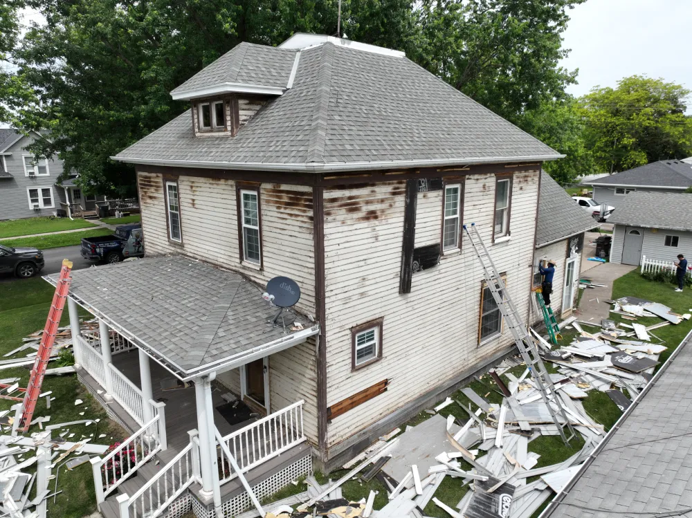 Old house during renovation. White siding is partially removed, revealing aged wood. Ladders surround the house. Debris litters the ground.