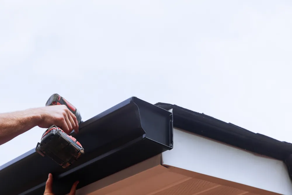 A person’s hand uses a power drill to attach a black rain gutter to the edge of a white roof under a clear sky. The scene conveys a DIY atmosphere.