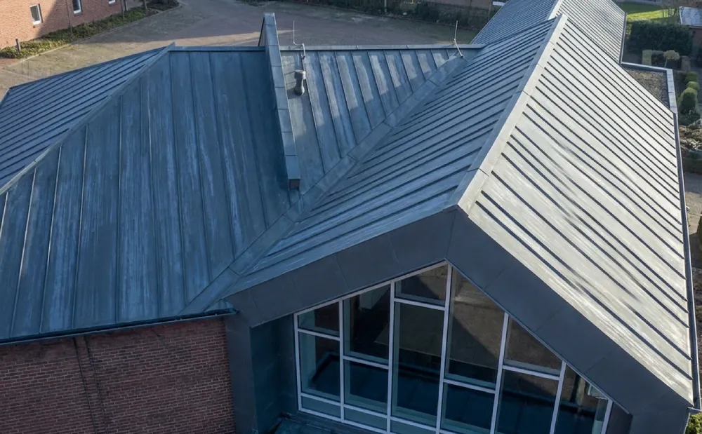 Aerial view of a building with a modern, sloped metal roof featuring triangular skylights. The structure has brick walls and large glass windows.