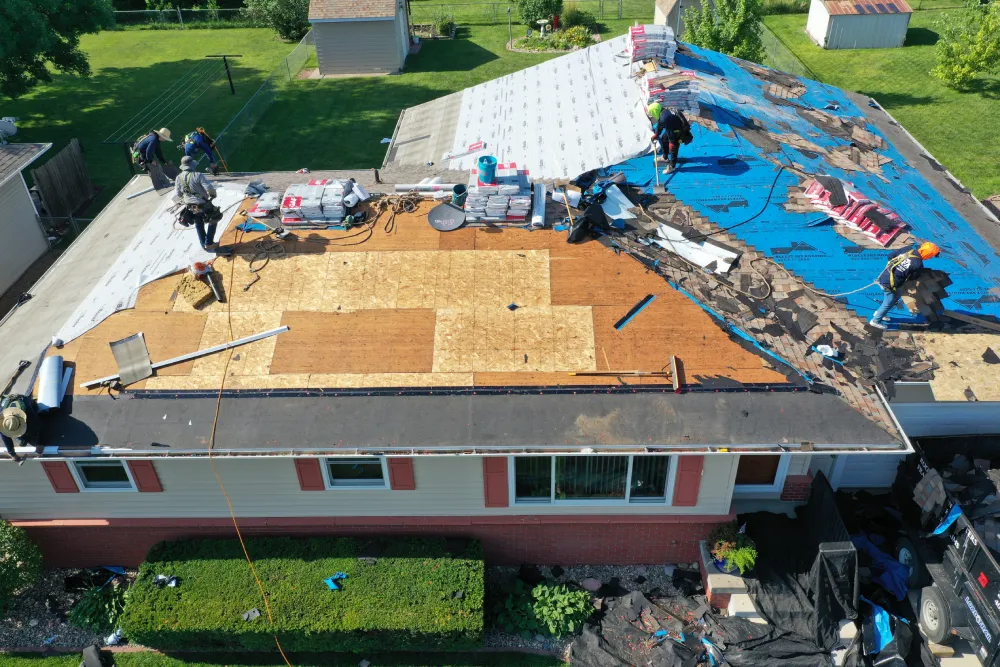 Aerial view of a house undergoing roof repairs. Workers in protective gear are laying new shingles, with blue tarps covering parts of the roof. Equipment and materials are scattered, indicating active work and progress. Surrounding green lawns add a calm contrast.