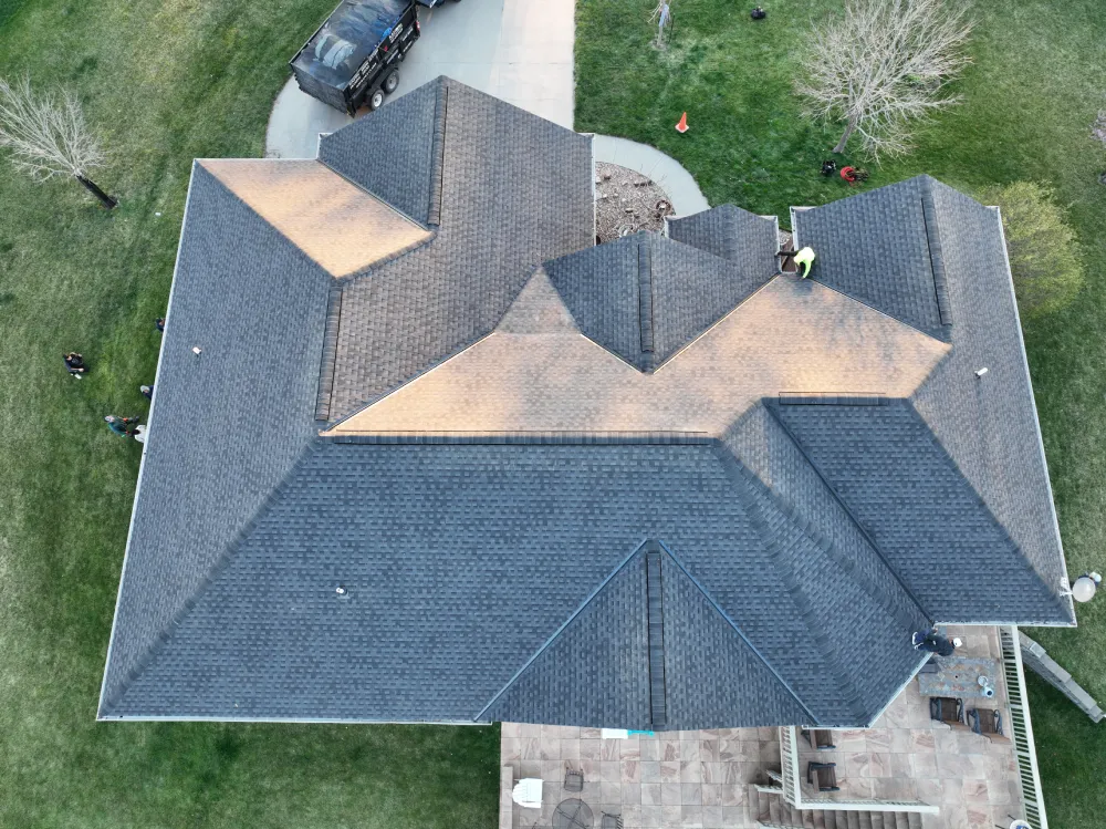 Aerial view of a large, complex roof with dark shingles, surrounded by green grass. A person works on the roof, and a truck is parked in the driveway.