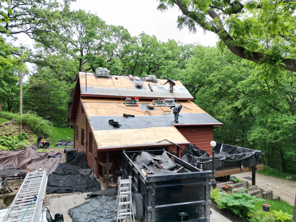 Roofers work on a large red house surrounded by lush green trees. Ladders and equipment are scattered around, conveying a busy, industrious scene.