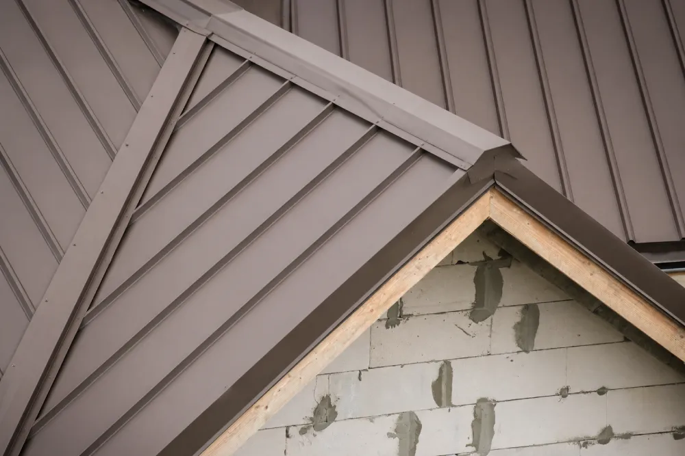 Close-up of a modern metal roof with interlocking panels. The sleek, gray-toned roof contrasts with the unfinished brick wall beneath, conveying a sense of construction.