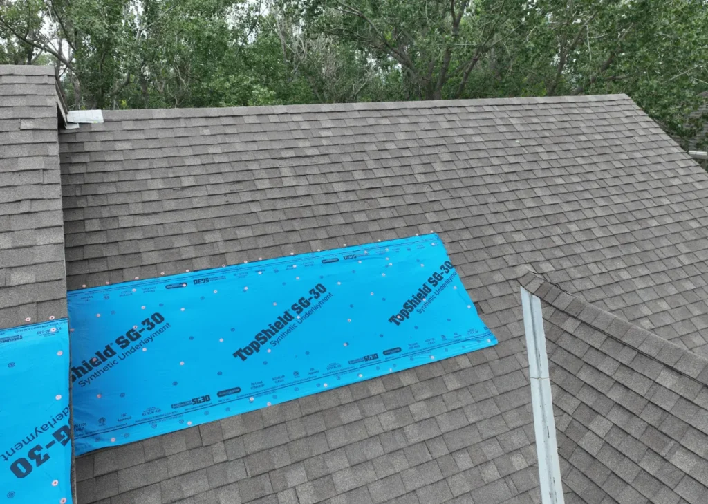 Gray shingled roof with a blue tarpaulin section labeled "TopShield SG-30" covering part of it, surrounded by green trees in the background.