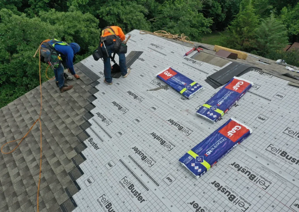 Two workers in safety gear install underlayment on a roof surrounded by trees. Roofing materials are scattered nearby, showcasing a construction scene.