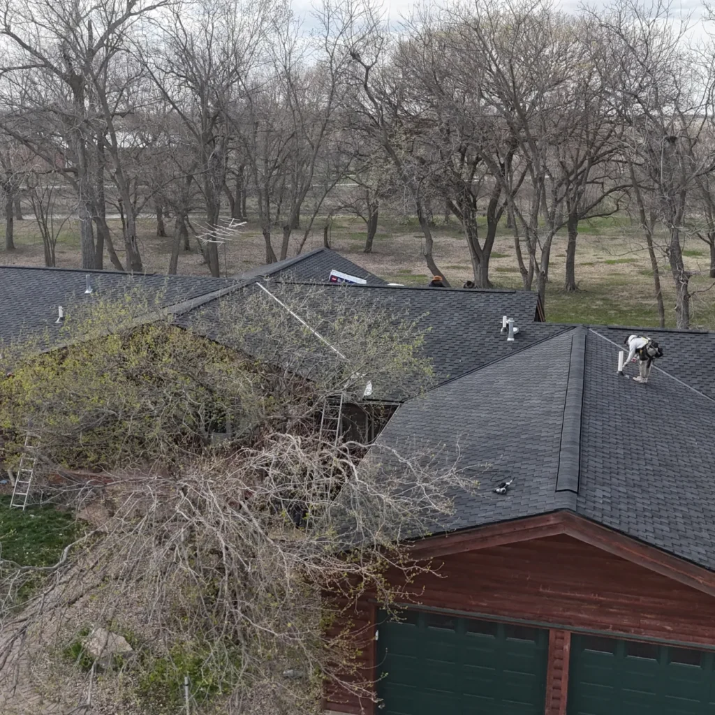 Aerial view of a large wooden house with a dark roof surrounded by leafless trees. People work on the roof, suggesting repair or maintenance tasks.